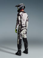 Zawodnik motocrossowy w pełnym stroju wyścigowym, koszulka 2026 Racer Lahnd i dopasowane spodnie w kolorze Sand Camo, szaro-czarny wzór kamuflażu z wyraźnymi logotypami Alpinestars, czarny kask, czarne buty i neonowożółte rękawice, widok z tyłu.