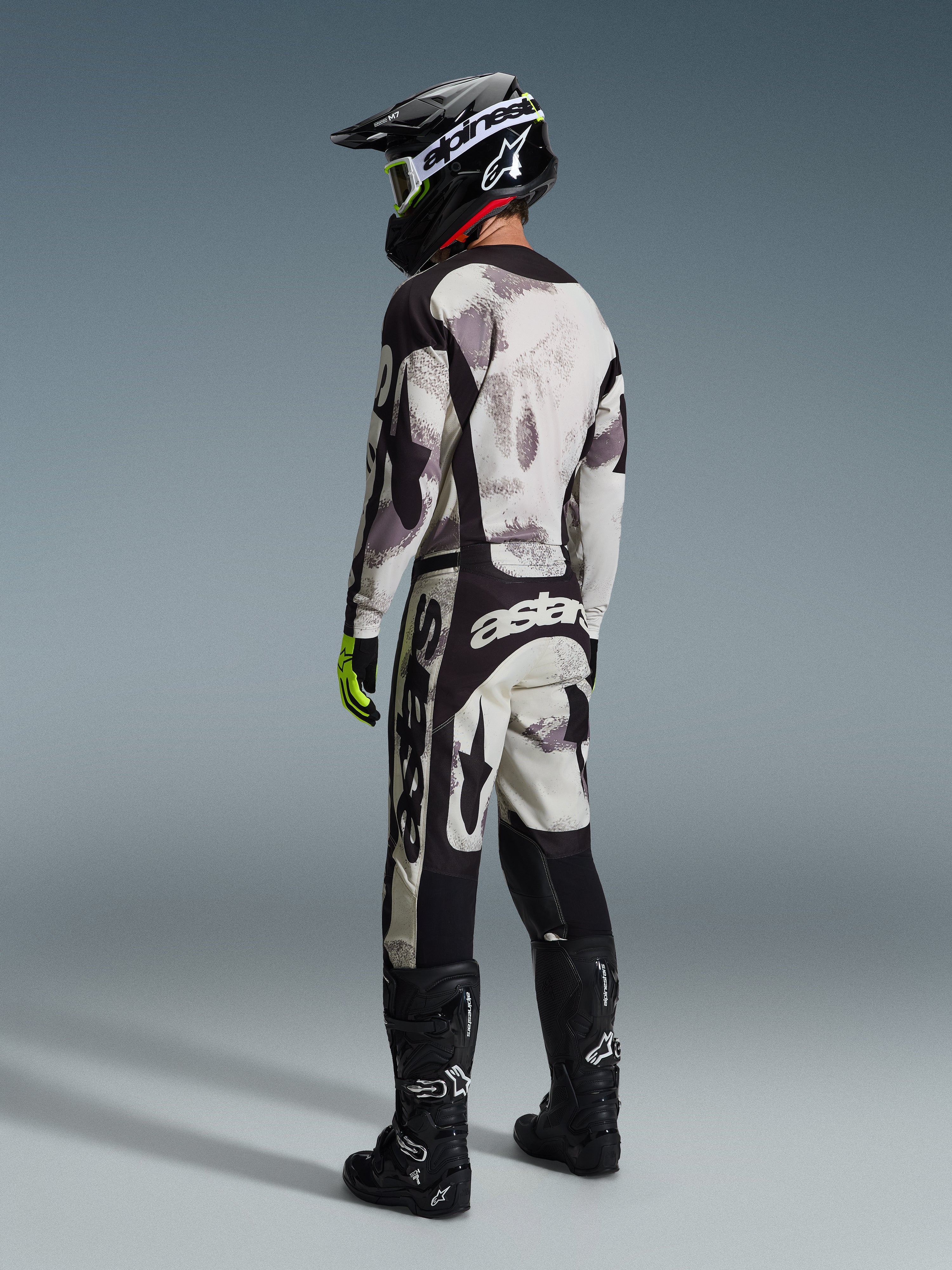 Zawodnik motocrossowy w pełnym stroju wyścigowym, koszulka 2026 Racer Lahnd i dopasowane spodnie w kolorze Sand Camo, szaro-czarny wzór kamuflażu z wyraźnymi logotypami Alpinestars, czarny kask, czarne buty i neonowożółte rękawice, widok z tyłu.