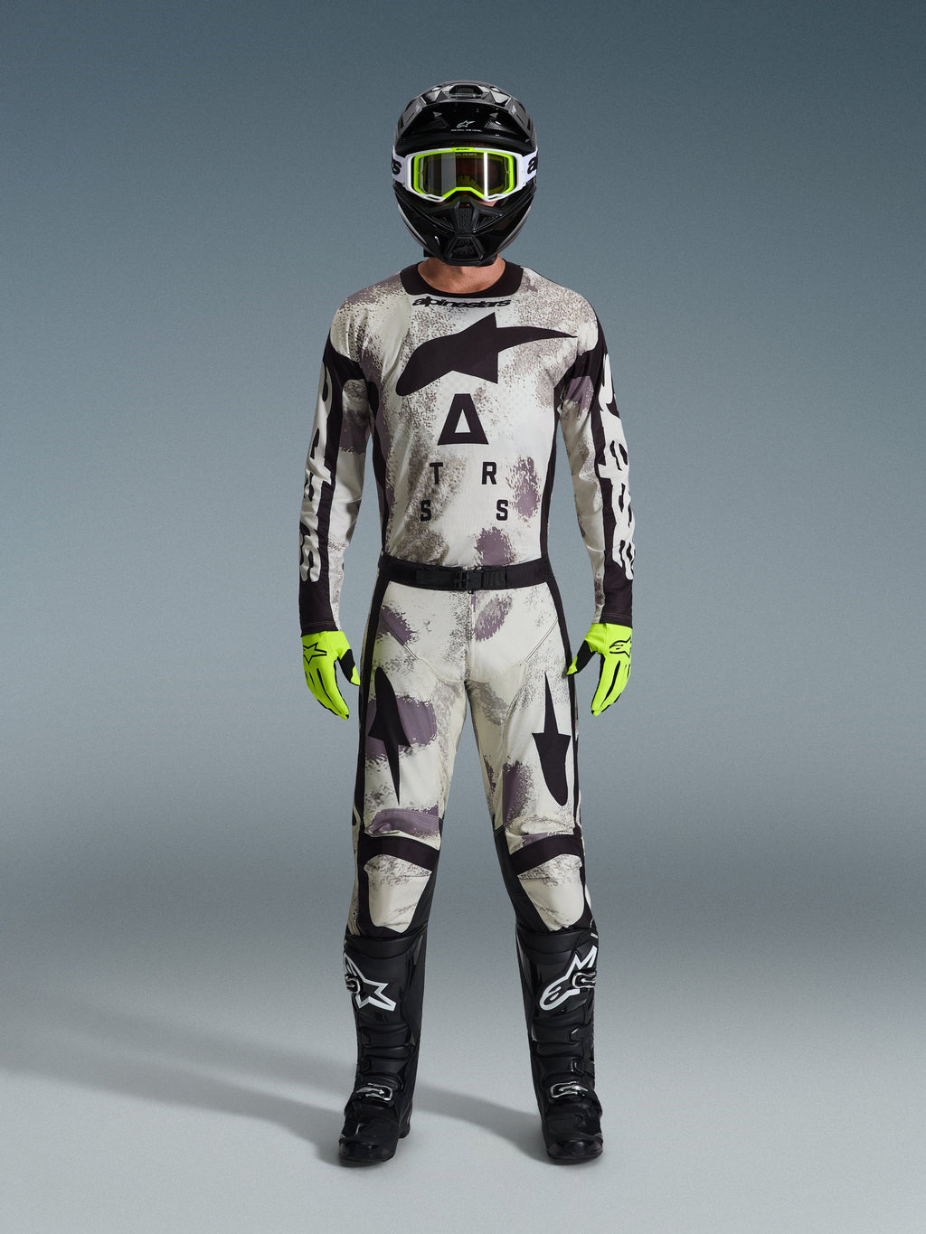 Motocyklista motocrossowy w pełnym stroju wyścigowym, spodnie Alpinestars 2026 Racer Lahnd i pasująca bluza w kolorze szarym Sand Camo, czarny kask z neonowożółtymi goglami, jaskrawe neonowożółte rękawice i czarne buty, profesjonalny zestaw do wyścigów off-road