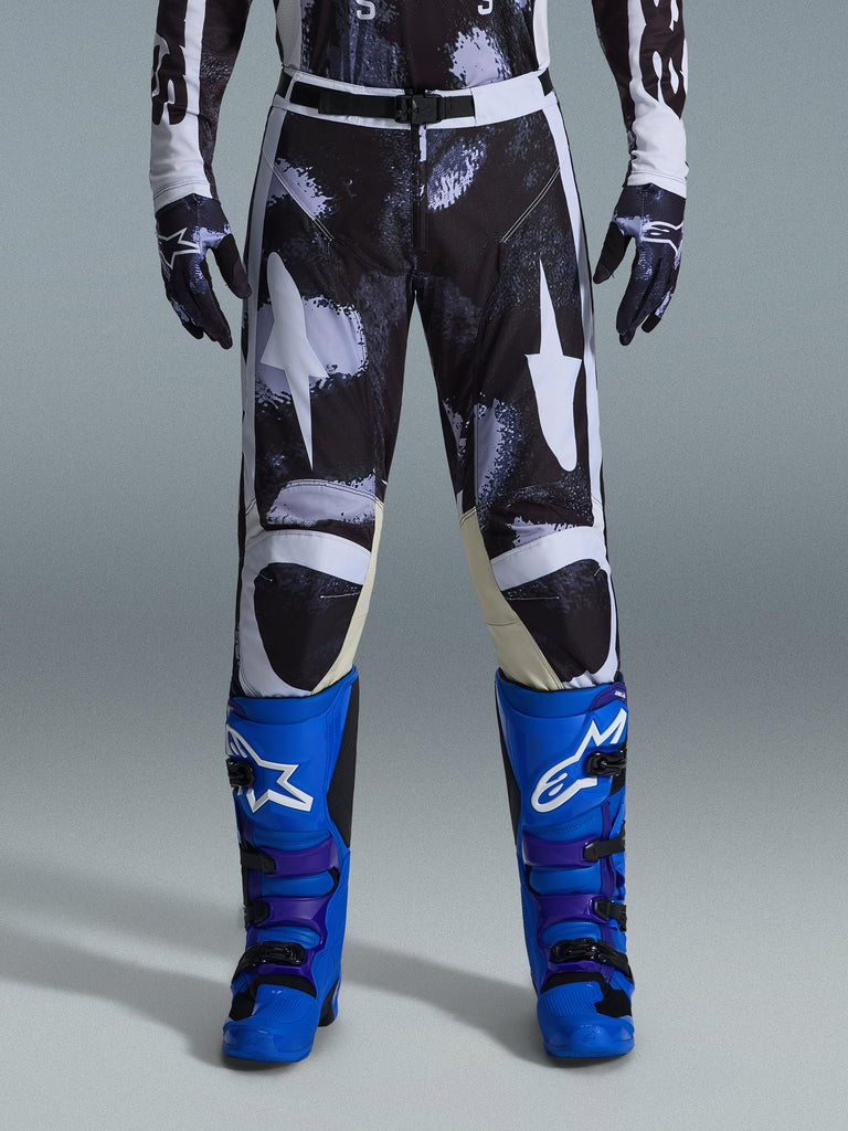 Alpinestars 2026 Racer Lahnd Pants, spodnie motocrossowe, Iron Camo, noszone z pasującymi rękawicami w kamuflażu oraz jaskrawoniebieskimi i fioletowymi butami, ze stylizowaną grafiką białych gwiazd na ciemnym wzorze moro, techniczne magnetyczne zapięcie, profesjonalna odzież do wyścigów off-road.