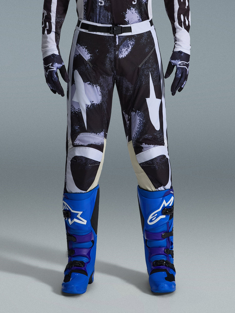 Alpinestars 2026 Racer Lahnd Pants, spodnie motocrossowe, Iron Camo, noszone z pasującymi rękawicami w kamuflażu oraz jaskrawoniebieskimi i fioletowymi butami, ze stylizowaną grafiką białych gwiazd na ciemnym wzorze moro, techniczne magnetyczne zapięcie, profesjonalna odzież do wyścigów off-road.