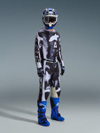 Motocyklista motocrossowy w pełnym rynsztunku, spodnie Alpinestars 2026 Racer Lahnd we wzorze Iron Camo i szarym, pasująca koszulka, jasnoniebieskie i fioletowe buty motocrossowe, niebiesko-biały kask, profesjonalny strój do wyścigów terenowych