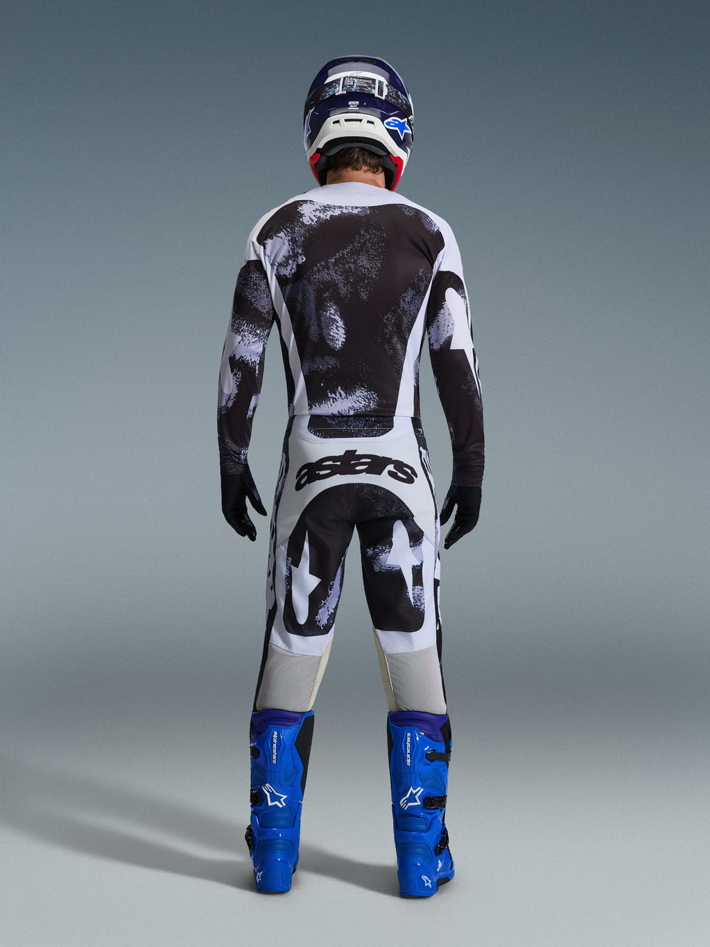 Alpinestars 2026 Racer Lahnd Pants, spodnie motocrossowe, szaro-czarny wzór Iron Camo, widok od tyłu motocyklisty w pełnym stroju wyścigowym z pasującą koszulką i niebieskimi butami, zaprojektowane z myślą o wydajności off-road