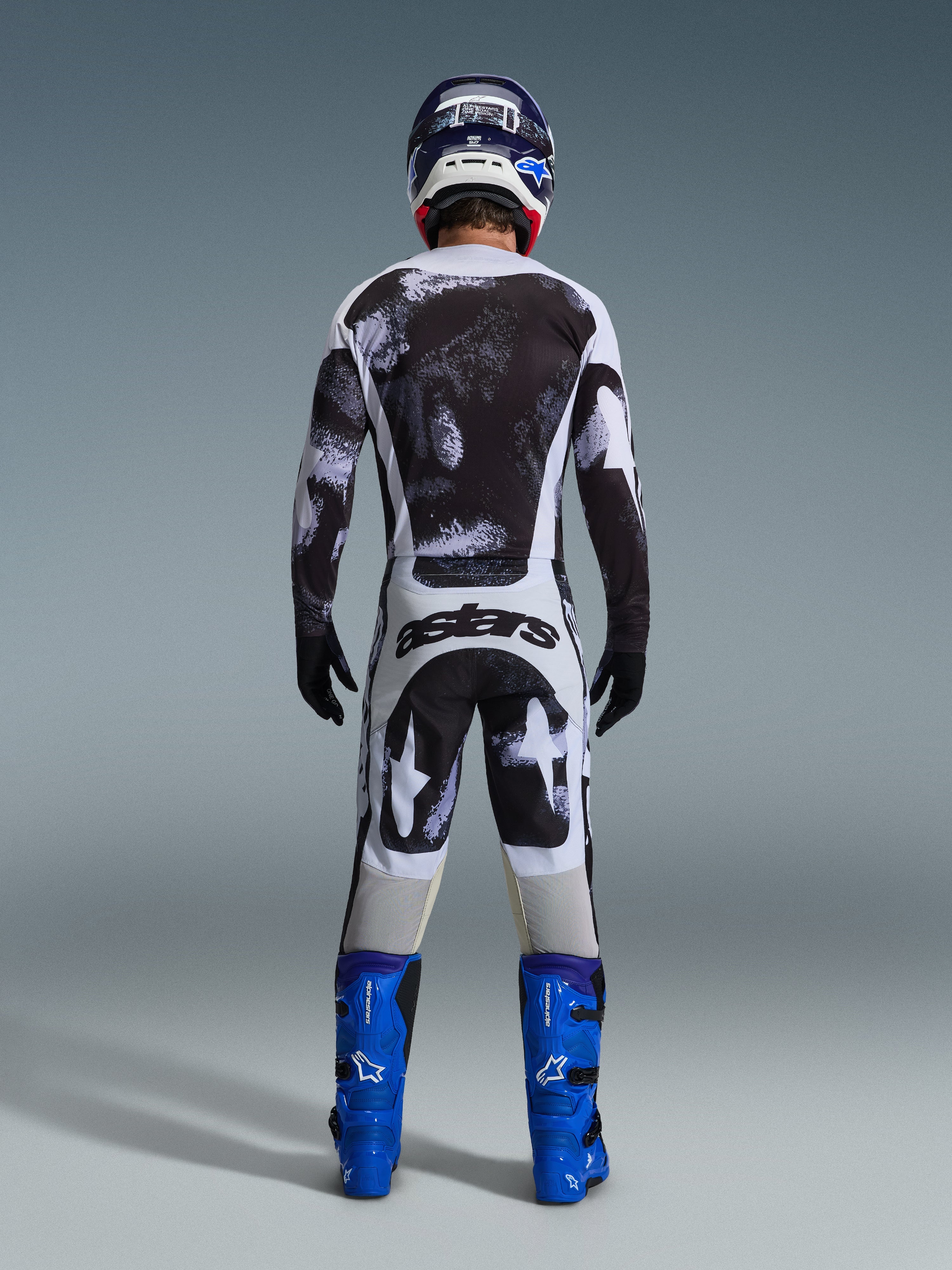 Alpinestars 2026 Racer Lahnd Pants, spodnie motocrossowe, szaro-czarny wzór Iron Camo, widok od tyłu motocyklisty w pełnym stroju wyścigowym z pasującą koszulką i niebieskimi butami, zaprojektowane z myślą o wydajności off-road
