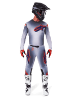 Zawodnik motocrossowy w spodniach Alpinestars Supertech Lipan 2025 i dopasowanej bluzie w kolorze szarym i pomarańczowym fluo, z wyraźnymi pomarańczowymi i granatowymi logotypami, w zestawie z czarnym kaskiem, czerwonymi rękawicami i biało-czarnymi butami, kompletny zestaw wyścigowy do jazdy terenowej.