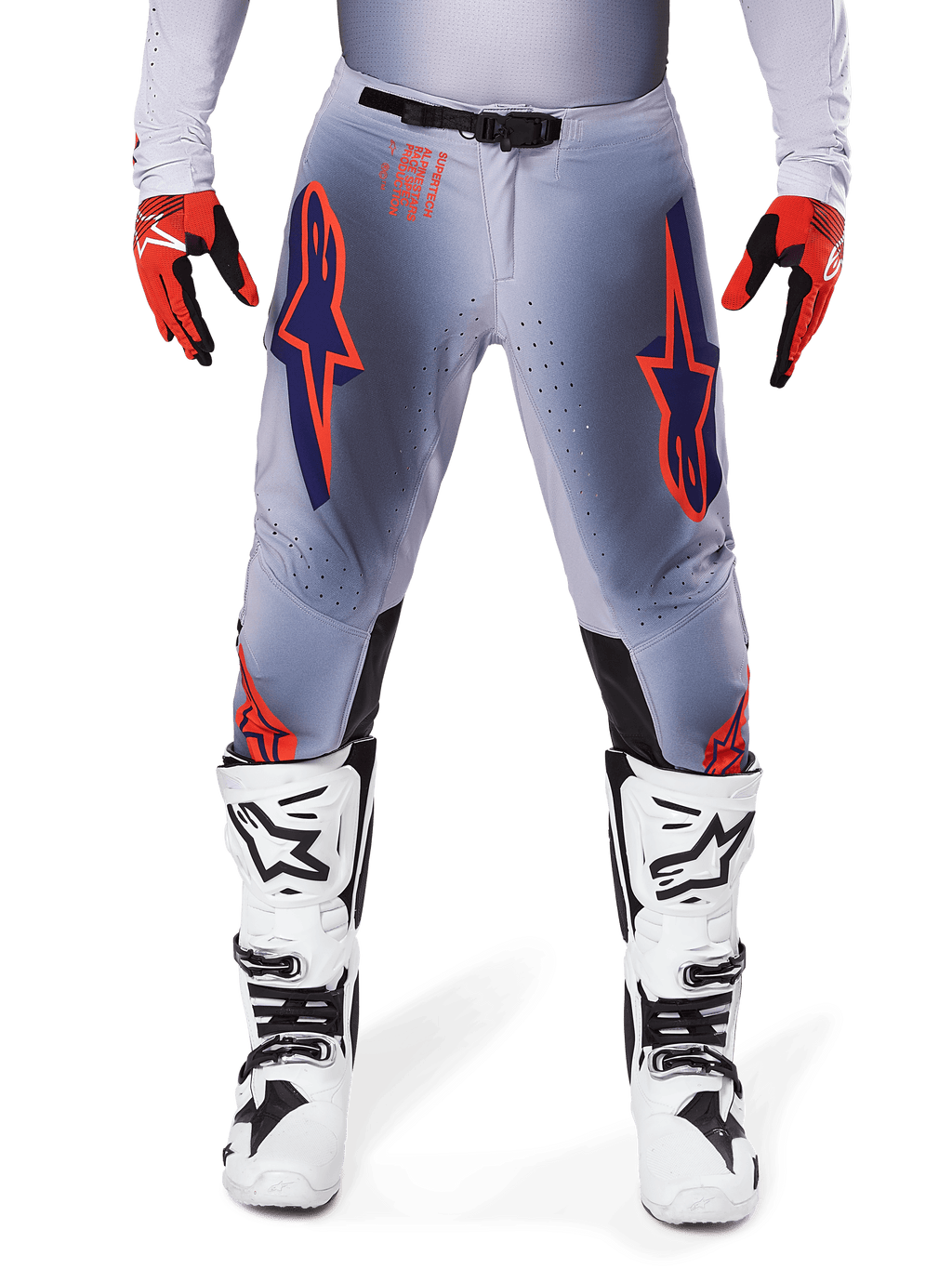 Spodnie Alpinestars Supertech Lipan 2025, spodnie motocrossowe, szaro-pomarańczowe fluo, zestawione z czerwono-czarnymi rękawicami i biało-czarnymi butami, z dużymi logotypami gwiazdy Alpinestars na udach oraz perforacjami wentylacyjnymi dla profesjonalnych zawodników.