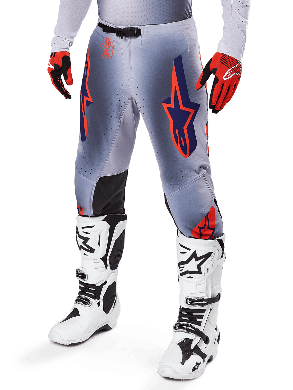 Alpinestars Supertech Lipan Pants 2025, spodnie motocrossowe, szare i pomarańczowe fluo, z granatowymi gwiazdami i czarnymi panelami wewnętrznymi na kolanach, noszone z czerwono-pomarańczowymi rękawicami i białymi butami, profesjonalna odzież do wyścigów terenowych