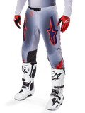 Alpinestars Supertech Lipan Pants 2025, spodnie motocrossowe, szare i pomarańczowe fluo, z granatowymi gwiazdami i czarnymi panelami wewnętrznymi na kolanach, noszone z czerwono-pomarańczowymi rękawicami i białymi butami, profesjonalna odzież do wyścigów terenowych
