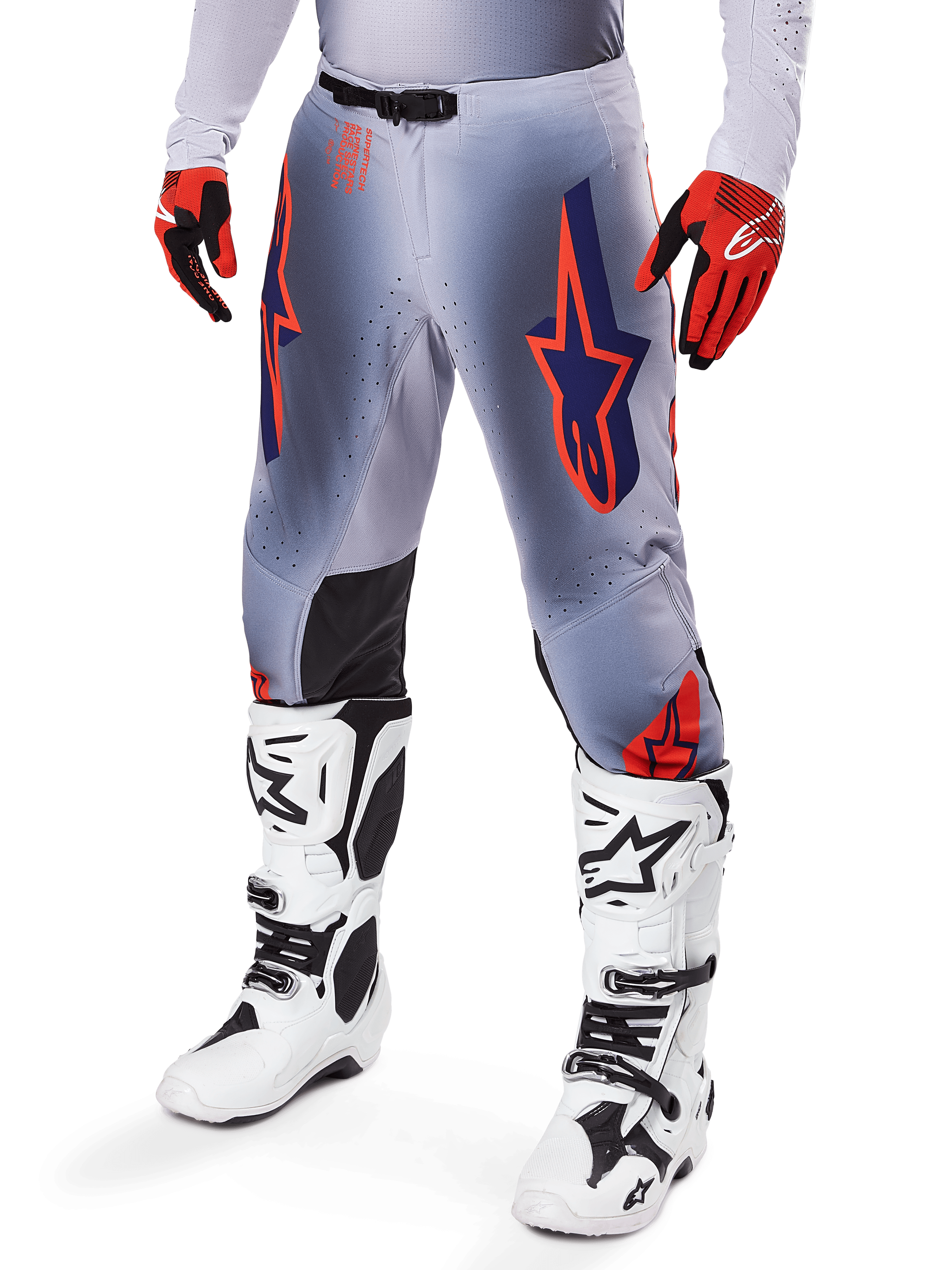Alpinestars Supertech Lipan Pants 2025, spodnie motocrossowe, szare i pomarańczowe fluo, z granatowymi gwiazdami i czarnymi panelami wewnętrznymi na kolanach, noszone z czerwono-pomarańczowymi rękawicami i białymi butami, profesjonalna odzież do wyścigów terenowych