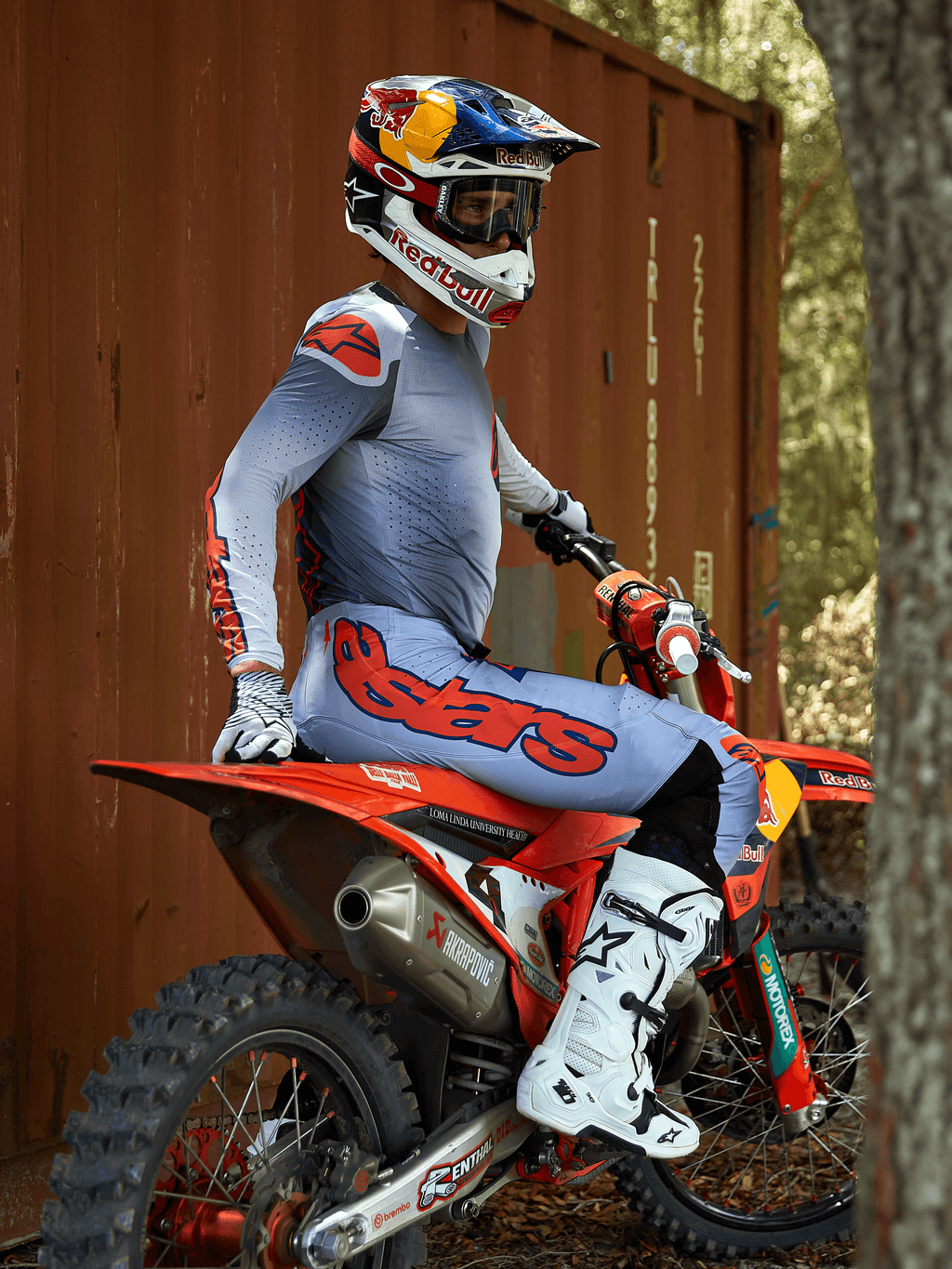 Motocyklista motocrossowy siedzący na czerwonym motocyklu crossowym, ubrany w zestaw odzieży Alpinestars 2025 Supertech Lipan w kolorze szarym i pomarańczowym fluo, jasnoniebieskie spodnie z wyraźnym pomarańczowym logo Astars, pasująca koszulka, białe buty motocrossowe oraz kask Red Bull, profesjonalny zestaw wyścigowy
