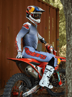 Motocyklista motocrossowy siedzący na czerwonym motocyklu crossowym, ubrany w zestaw odzieży Alpinestars 2025 Supertech Lipan w kolorze szarym i pomarańczowym fluo, jasnoniebieskie spodnie z wyraźnym pomarańczowym logo Astars, pasująca koszulka, białe buty motocrossowe oraz kask Red Bull, profesjonalny zestaw wyścigowy