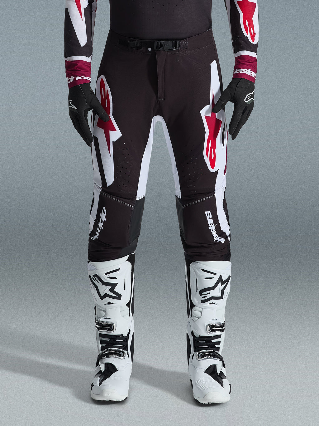 Alpinestars 2026 Supertech AFD Vista Pants, spodnie motocrossowe, czarno-czerwono-białe, z dużymi graficznymi logotypami na udach i kolanach, noszone z białymi butami wyścigowymi i czarnymi rękawicami, sportowy design gotowy do wyścigów dla elity zawodów off-road
