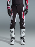 Alpinestars 2026 Supertech AFD Vista Pants, spodnie motocrossowe, czarno-czerwono-białe, z dużymi graficznymi logotypami na udach i kolanach, noszone z białymi butami wyścigowymi i czarnymi rękawicami, sportowy design gotowy do wyścigów dla elity zawodów off-road