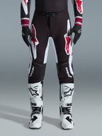Alpinestars 2026 Supertech AFD Vista Pants, spodnie motocrossowe, czarno-czerwono-białe, z dużymi graficznymi logotypami na udach i kolanach, noszone z białymi butami wyścigowymi i czarnymi rękawicami, sportowy design gotowy do wyścigów dla elity zawodów off-road