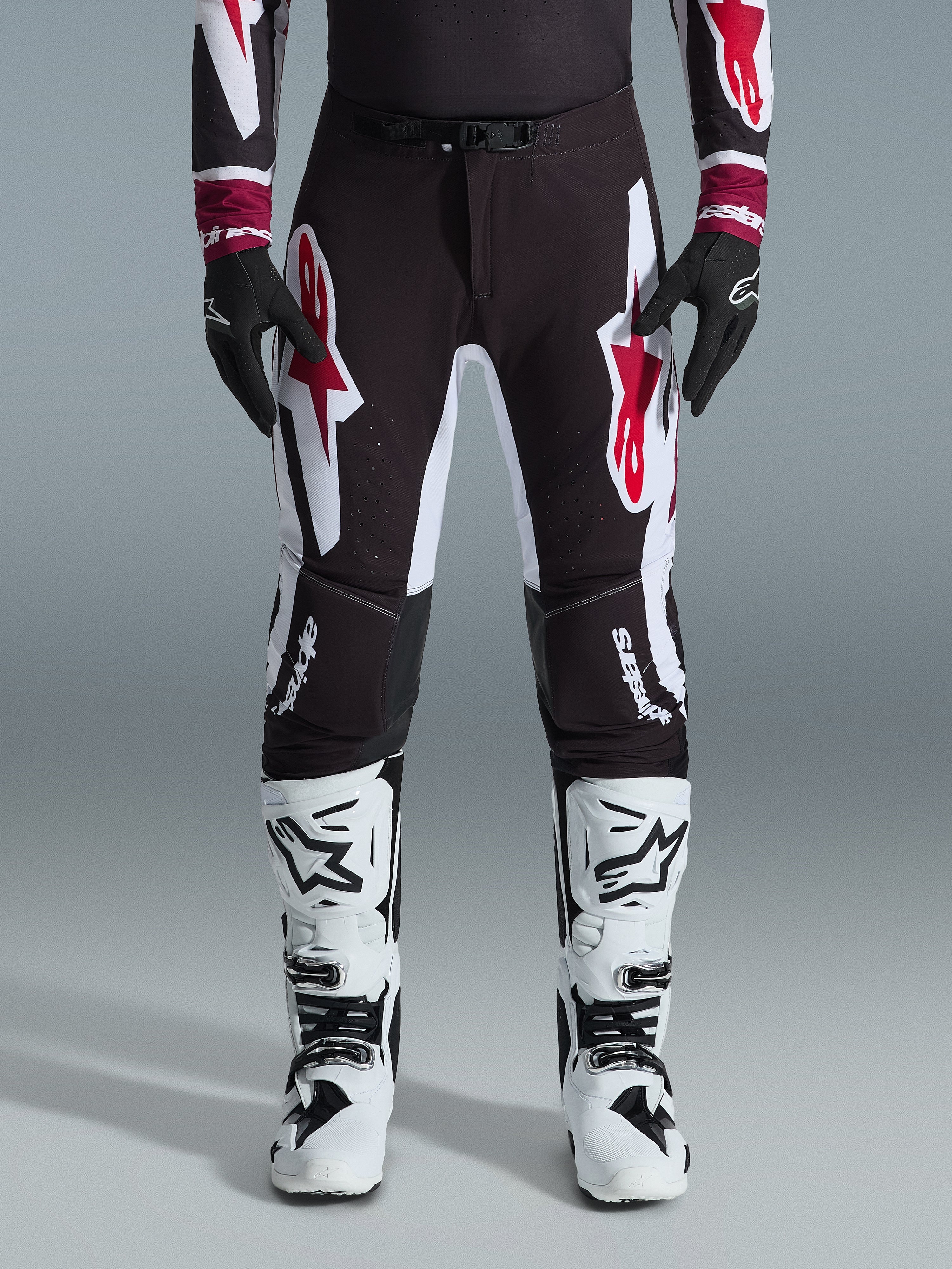 Alpinestars 2026 Supertech AFD Vista Pants, spodnie motocrossowe, czarno-czerwono-białe, z dużymi graficznymi logotypami na udach i kolanach, noszone z białymi butami wyścigowymi i czarnymi rękawicami, sportowy design gotowy do wyścigów dla elity zawodów off-road