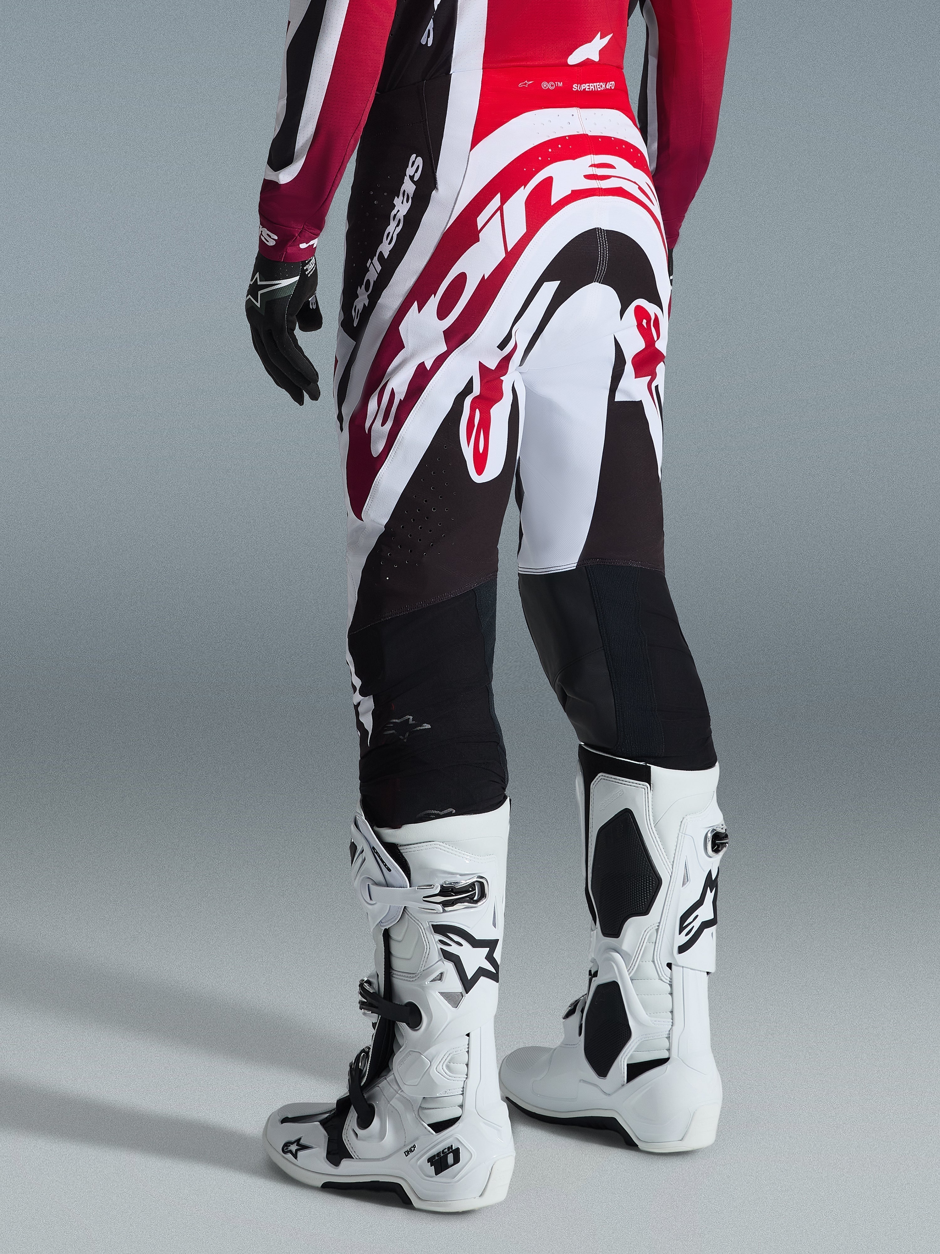 Alpinestars 2026 Supertech AFD Vista Pants, spodnie motocrossowe, czarno-czerwono-białe, widok z tyłu z dużym logo Alpinestars na siedzeniu, zestawione z białymi butami Tech 10 i czarnymi rękawicami, sportowy design dla elitarnej wydajności off-road