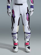 Alpinestars 2026 Supertech AFD Vista Pants, spodnie motocrossowe, białe, fioletowe i czarne, noszone z pasującymi białymi butami i rękawicami, z logo Astars z gradientem od różu do fioletu na udach, profesjonalny design wyścigowy zapewniający wydajność w terenie.