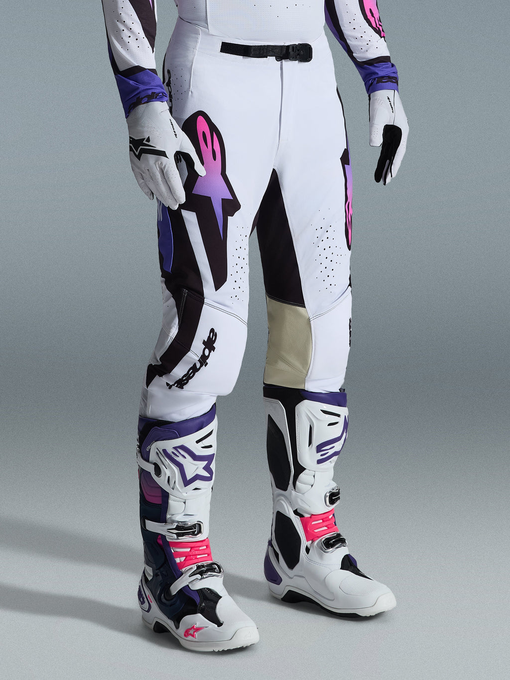 Alpinestars 2026 Supertech AFD Vista Pants, spodnie motocrossowe, białe, fioletowe i czarne, z różowo-fioletowym gradientowym logo Astar, noszone z pasującymi białymi i fioletowymi butami oraz rękawicami motocrossowymi, sportowy krój wyścigowy z beżowymi skórzanymi panelami po wewnętrznej stronie kolan i perforowanymi detalami.