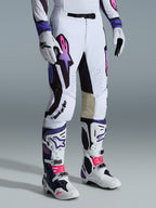 Alpinestars 2026 Supertech AFD Vista Pants, spodnie motocrossowe, białe, fioletowe i czarne, z różowo-fioletowym gradientowym logo Astar, noszone z pasującymi białymi i fioletowymi butami oraz rękawicami motocrossowymi, sportowy krój wyścigowy z beżowymi skórzanymi panelami po wewnętrznej stronie kolan i perforowanymi detalami.