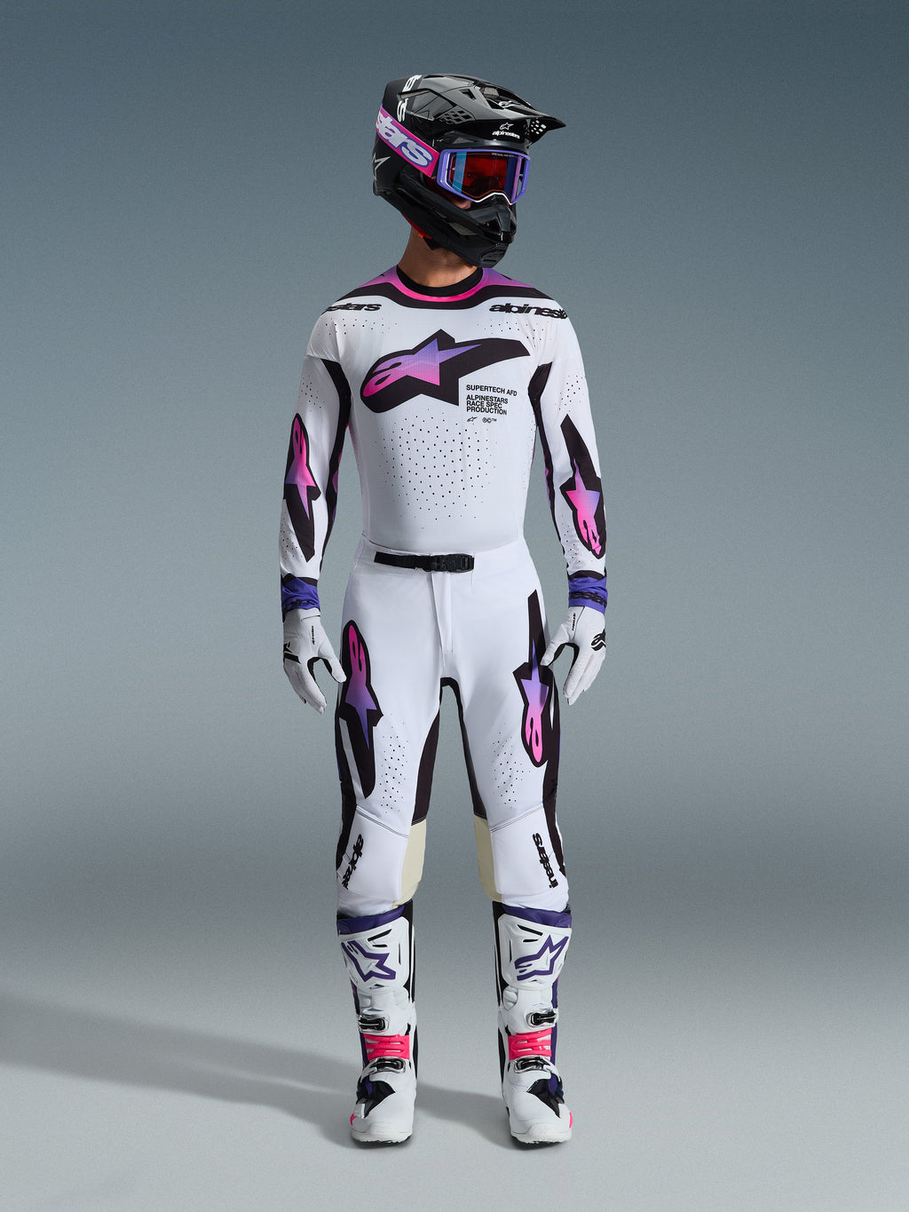 Motocyklista motocrossowy w kompletnym zestawie wyścigowym, spodnie i koszulka Alpinestars 2026 Supertech AFD Vista w kolorach białym, fioletowym i czarnym, dopasowane rękawice, biało-czarne buty, czarny kask z fioletowymi goglami, sportowy krój zapewniający wydajność w terenie