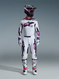 Zawodnik motocrossowy w zestawie Alpinestars 2026 Supertech AFD Vista, biała koszulka i spodnie z różowo-fioletowymi grafikami gwiazd z gradientem, czarny kask z dopasowanymi goglami, białe buty z różowymi akcentami i białe rękawice, profesjonalny strój do wyścigów off-road