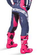 Alpinestars Techstar Dreem Pants 2025, spodnie motocrossowe, szare i jaskraworóżowe, noszone z pasującymi różowymi butami, z dużym białym napisem Alpinestars na siedzeniu i logo gwiazdy na nogawkach, wytrzymały sportowy krój wyścigowy do jazdy w terenie