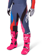 Alpinestars Techstar Dreem Pants 2025, spodnie motocrossowe, ciemnoszare z niebieskimi, czerwonymi i różowymi panelami, białe grafiki, noszone z jaskraworóżowymi butami i czarno-białymi rękawicami, wyrazisty design o wysokiej widoczności