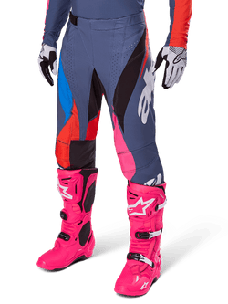 Alpinestars Techstar Dreem Pants 2025, spodnie motocrossowe, ciemnoszare z niebieskimi, czerwonymi i różowymi panelami, białe grafiki, noszone z jaskraworóżowymi butami i czarno-białymi rękawicami, wyrazisty design o wysokiej widoczności