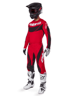 Motocyklista motocrossowy w kompletnym stroju wyścigowym Alpinestars, spodnie Techstar Dreem 2025 w kolorze jasnoczerwonym i czarnym, pasująca czerwono-czarna koszulka z białym logo, czarny kask oraz biało-czarne buty, skoordynowany zestaw do wyścigów terenowych