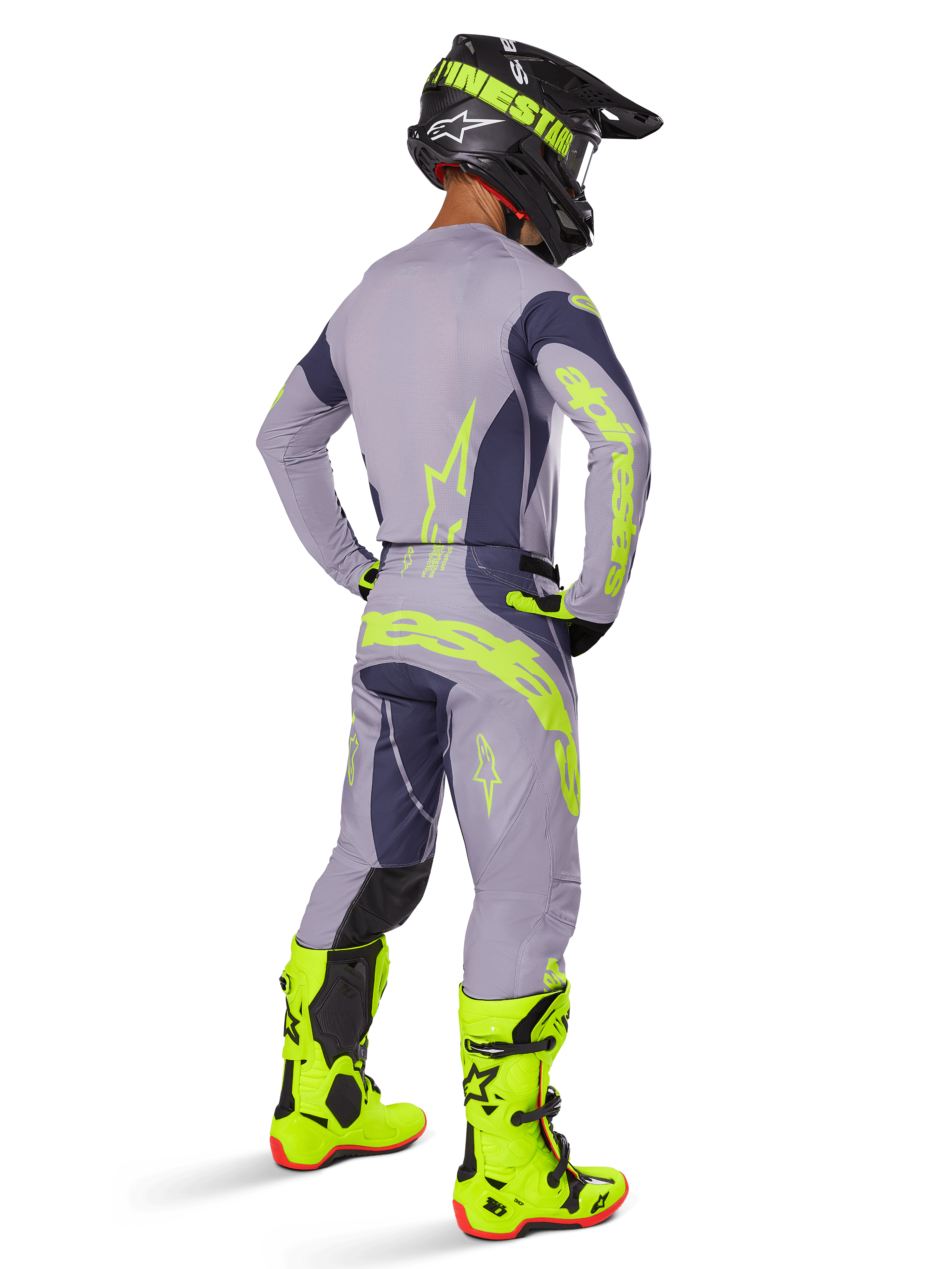 Zawodnik motocrossowy widziany od tyłu w pełnym stroju wyścigowym, w tym spodnie Alpinestars 2025 Techstar Dreem w kolorze szarym i ciemnoszarym z wyraźnymi neonowożółtymi akcentami, dopasowana szara bluza z neonowymi grafikami, jaskrawe neonowożółte buty i czarny kask; profesjonalny, spójny zestaw motocrossowy.