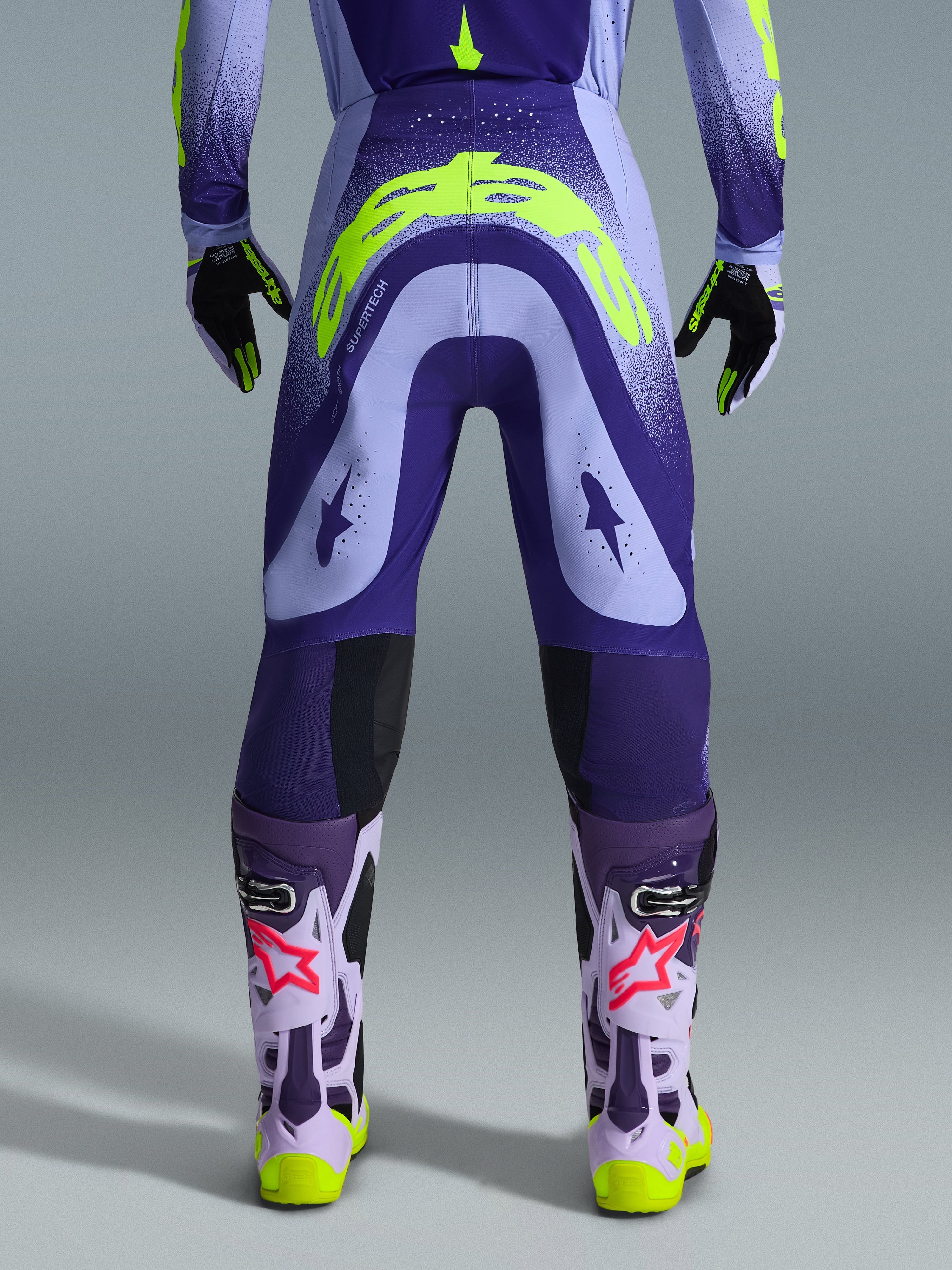 Alpinestars 2026 Supertech Scenz Pants, spodnie motocrossowe, fioletowe i neonowożółte (fluo), widok z tyłu pokazujący duże żółte logo na siedzeniu, w zestawie z pasującą bluzą, rękawicami i butami ochronnymi, sportowy krój do profesjonalnych wyścigów motocrossowych