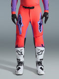 Alpinestars 2026 Supertech Scenz Pants, spodnie motocrossowe, kolor koralowy i fioletowy, noszone z dopasowaną koszulką, biało-czarnymi butami i czarnymi rękawicami, duże fioletowe logo Astars na udach, czarna magnetyczna klamra w pasie, profesjonalny krój sportowy do wyścigów