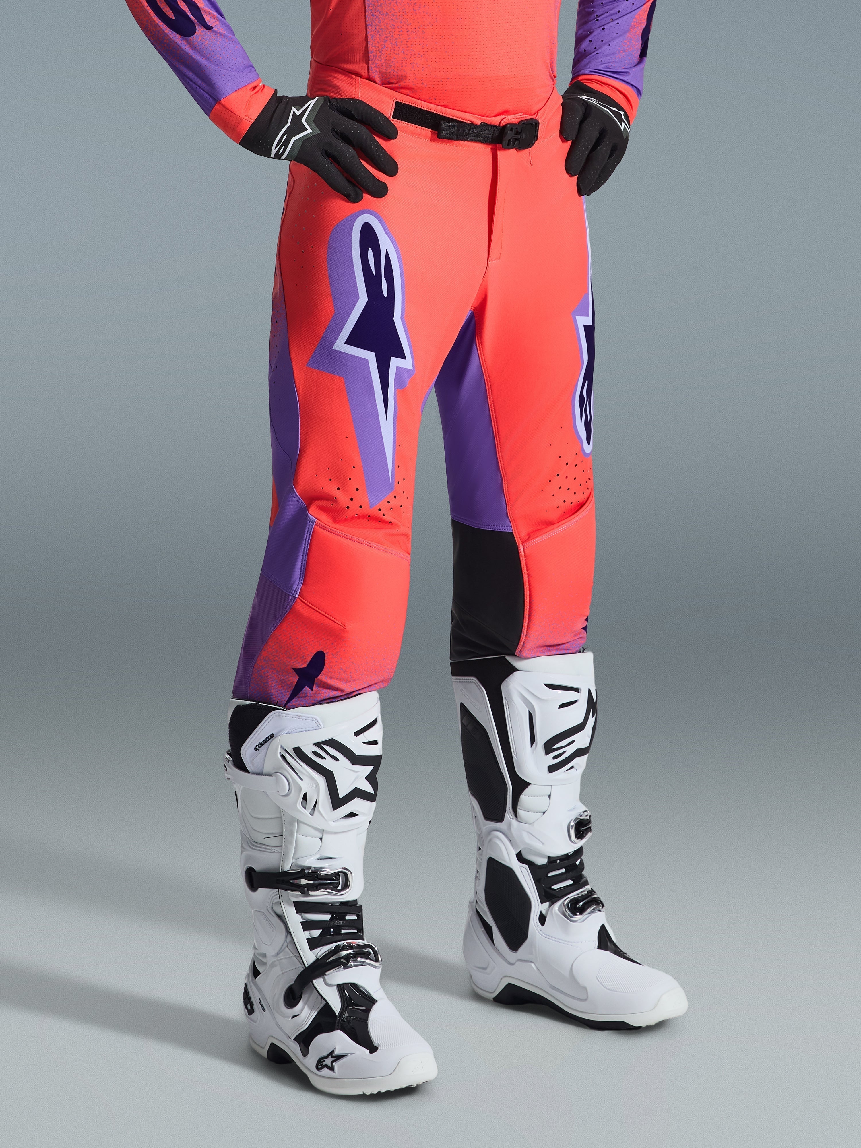 Alpinestars 2026 Supertech Scenz Pants, spodnie motocrossowe, kolor jaskrawy koralowy i fioletowy, noszone z biało-czarnymi butami motocrossowymi i czarnymi rękawicami, duże fioletowe logo Alpinestars, czarne skórzane panele na wewnętrznej stronie kolan, sportowy krój wyścigowy