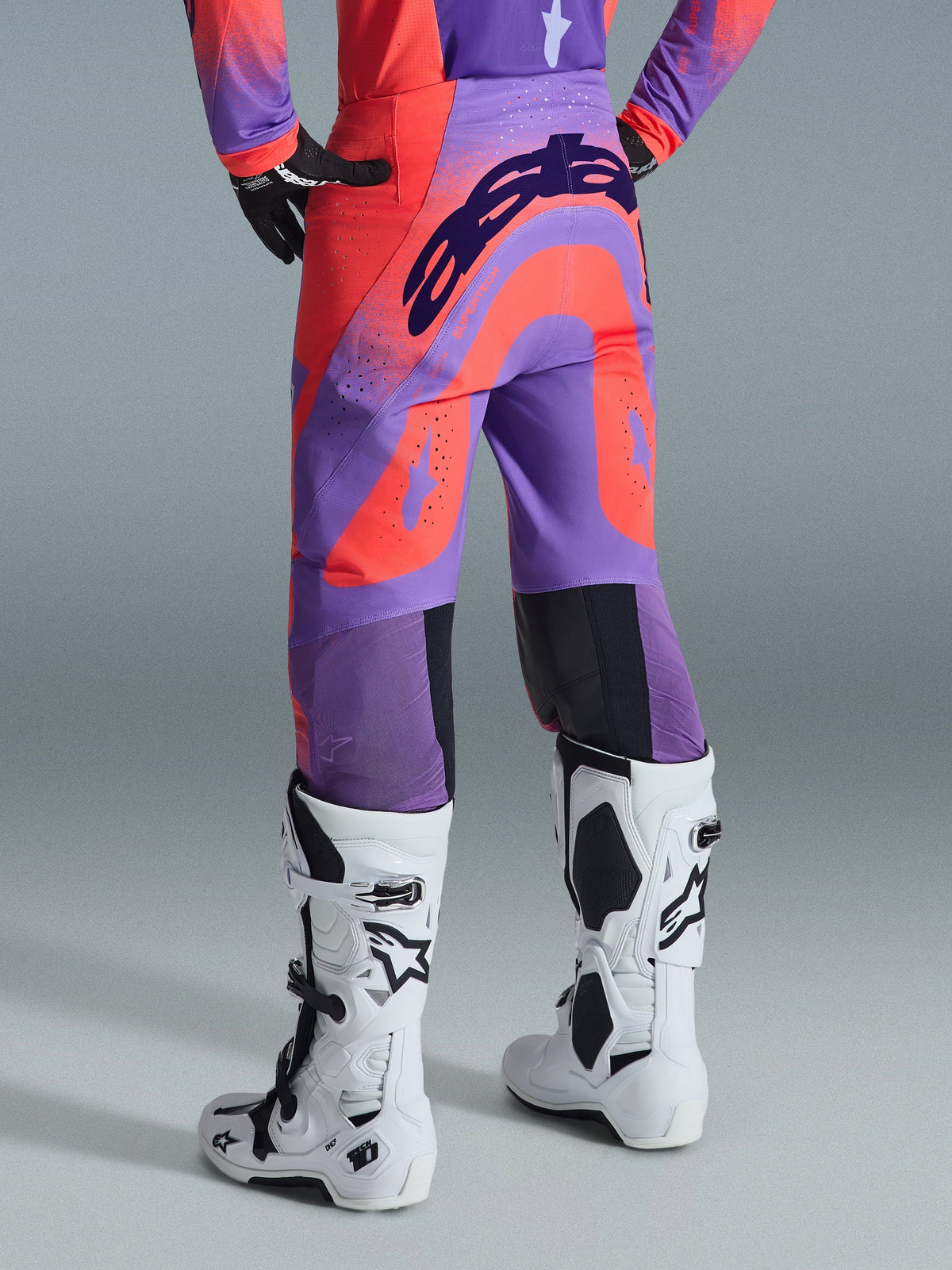 Alpinestars 2026 Supertech Scenz Pants, spodnie motocrossowe, kolor Hot Coral Purple, widok z tyłu z wyrazistym brandingiem na siedzisku, jaskrawe koralowe i fioletowe panele z perforowanymi strefami dla przepływu powietrza, zestawione z biało-czarnymi butami wyścigowymi