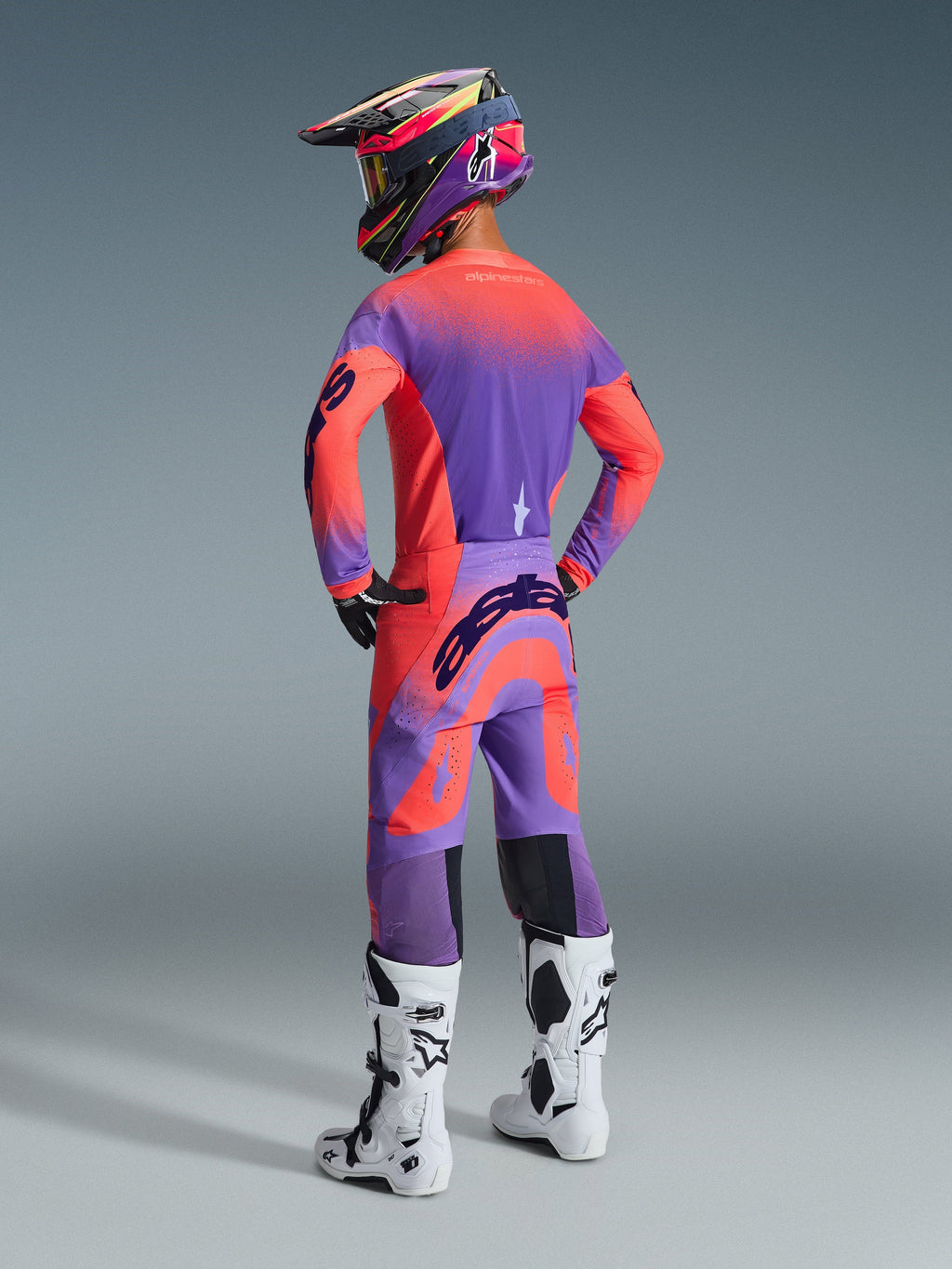 Alpinestars 2026 Supertech Scenz Pants, spodnie motocrossowe, kolor koralowy i fioletowy, widok z tyłu ukazujący wyraźne logo na siedzisku, noszone przez zawodnika z pasującą bluzą i białymi butami, profesjonalna odzież do wyścigów off-road.