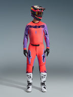 Alpinestars 2026 Supertech Scenz Pants, spodnie motocrossowe, kolor Hot Coral i fioletowy, noszone jako kompletny zestaw z pasującą koszulką i kaskiem, biało-czarne buty, fioletowe logotypy i sportowy krój wyścigowy.