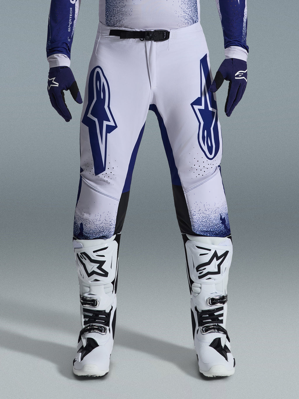 Alpinestars 2026 Supertech Scenz Pants, spodnie motocrossowe, jasnoszare i ciemnoniebieskie, noszone z biało-czarnymi butami motocrossowymi i ciemnoniebieskimi rękawicami, z dużymi niebieskimi logo Alpinestars na udach, magnetyczną klamrą SX1 i sportowym krojem do wyścigów terenowych