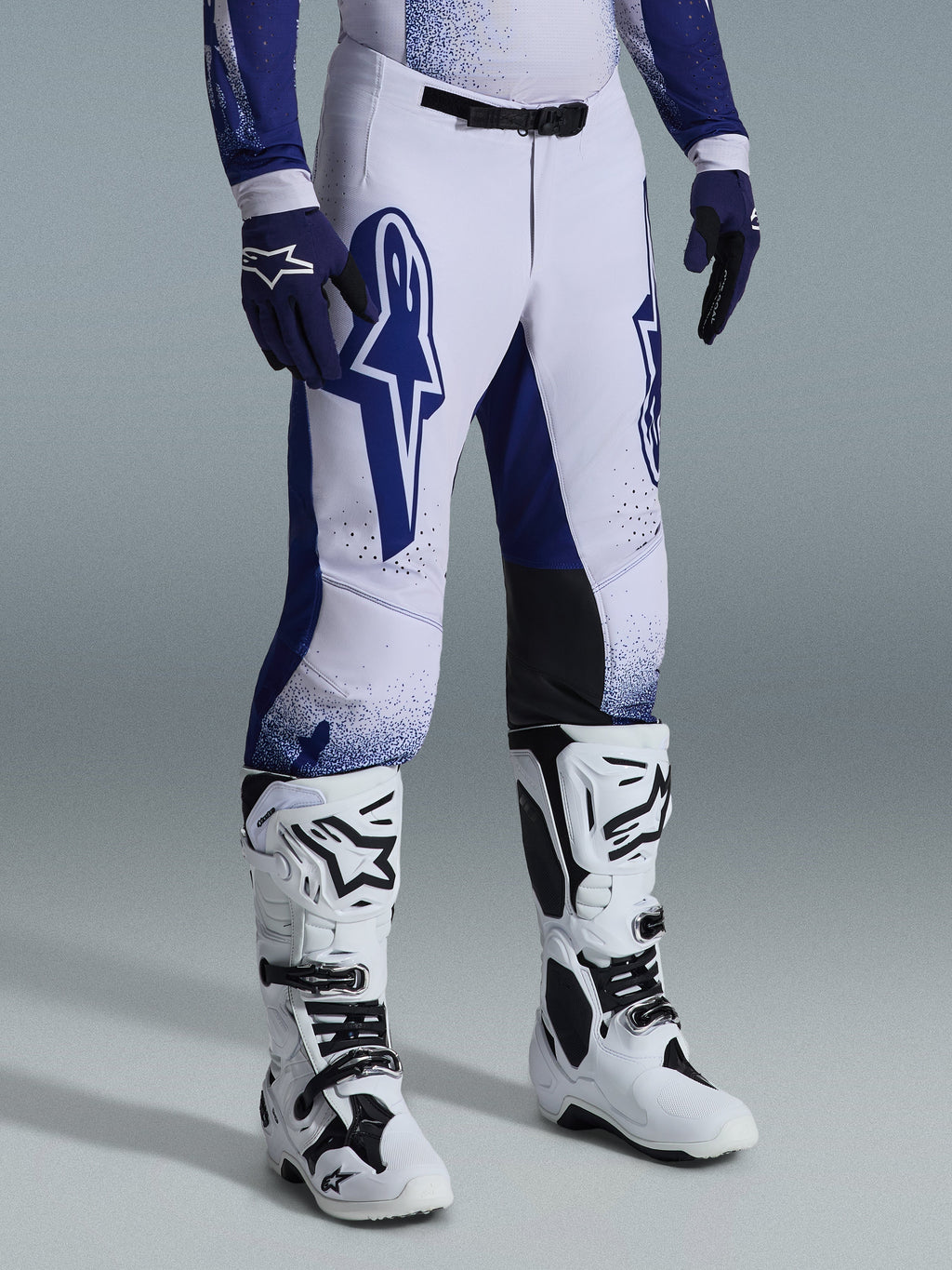 Alpinestars Supertech Scenz Pants 2026, spodnie motocrossowe, jasnoszare i ciemnoniebieskie, noszone z białymi butami motocrossowymi i niebieskimi rękawicami, duże niebieskie logo Alpinestars na udach, czarne panele na kolanach i kropkowany wzór graficzny, profesjonalny sprzęt do wyścigów off-road