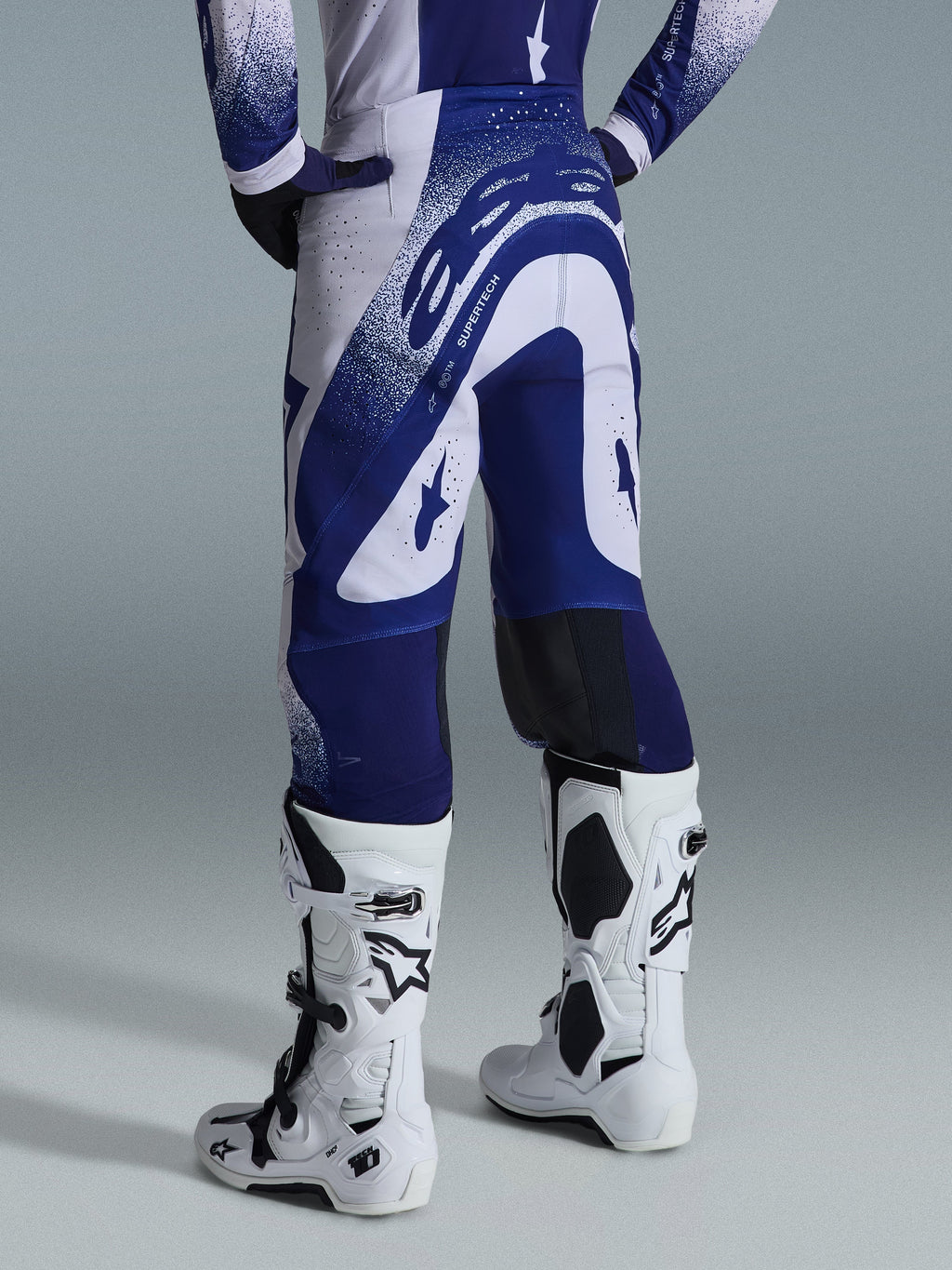 Alpinestars Supertech Scenz Pants 2026, spodnie motocrossowe, jasnoszare i ciemnoniebieskie, widok z tyłu w zestawie z pasującą bluzą i białymi butami, wyrazista niebieska grafika z nakrapianymi akcentami, sportowy krój wyczynowy do wyścigów off-road