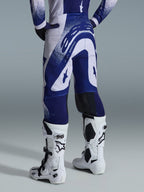 Alpinestars Supertech Scenz Pants 2026, spodnie motocrossowe, jasnoszare i ciemnoniebieskie, widok z tyłu w zestawie z pasującą bluzą i białymi butami, wyrazista niebieska grafika z nakrapianymi akcentami, sportowy krój wyczynowy do wyścigów off-road
