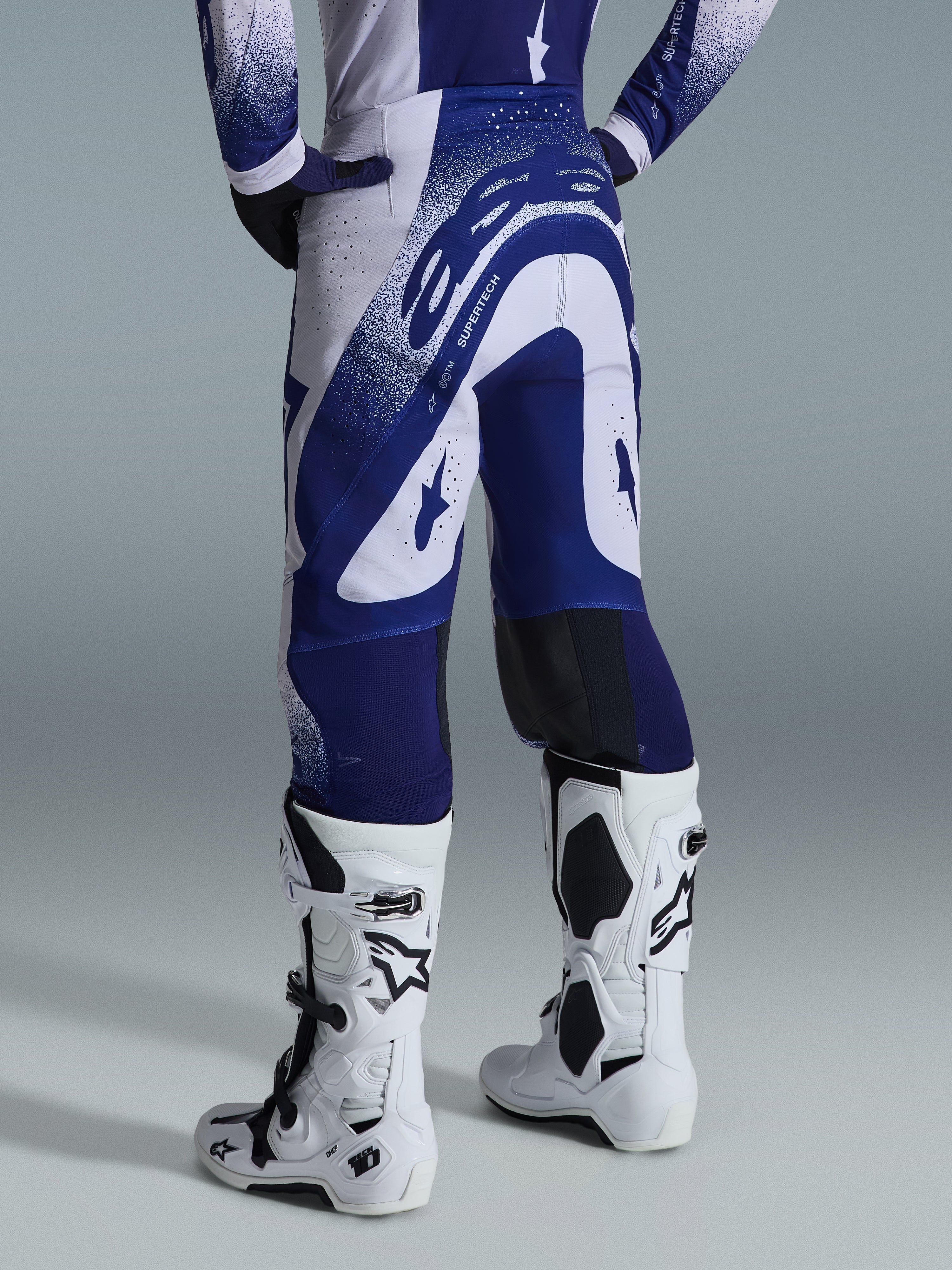 Alpinestars Supertech Scenz Pants 2026, spodnie motocrossowe, jasnoszare i ciemnoniebieskie, widok z tyłu w zestawie z pasującą bluzą i białymi butami, wyrazista niebieska grafika z nakrapianymi akcentami, sportowy krój wyczynowy do wyścigów off-road
