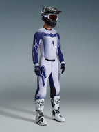 Motocyklista motocrossowy w spodniach 2026 Supertech Scenz w kolorze jasnoszarym i ciemnoniebieskim, dopasowana niebiesko-biała bluza z logo „astars” i efektem sprayu, czarny kask z białymi goglami, biało-czarne buty motocrossowe, profesjonalny zestaw do wyścigów terenowych.