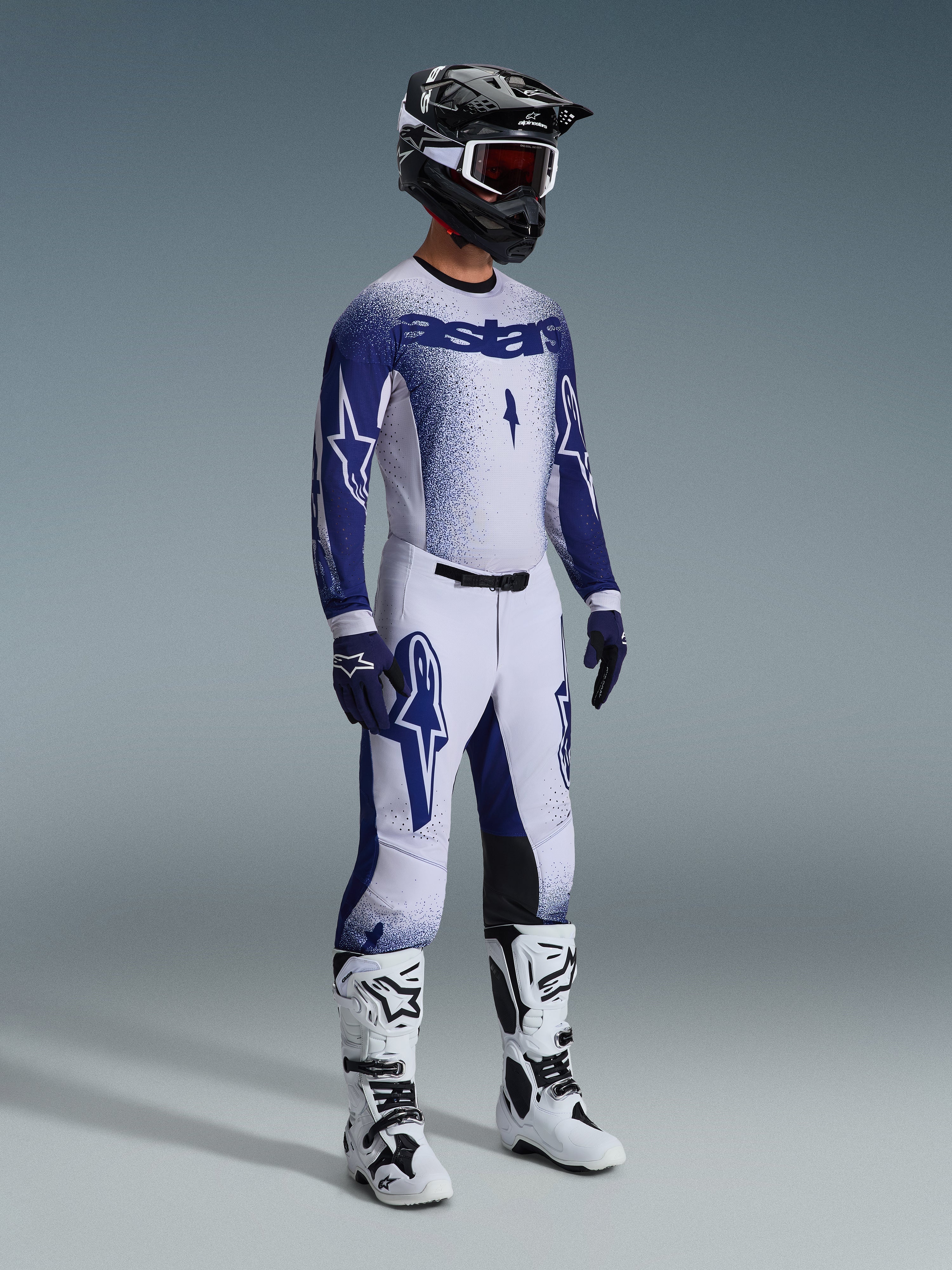 Motocyklista motocrossowy w spodniach 2026 Supertech Scenz w kolorze jasnoszarym i ciemnoniebieskim, dopasowana niebiesko-biała bluza z logo „astars” i efektem sprayu, czarny kask z białymi goglami, biało-czarne buty motocrossowe, profesjonalny zestaw do wyścigów terenowych.