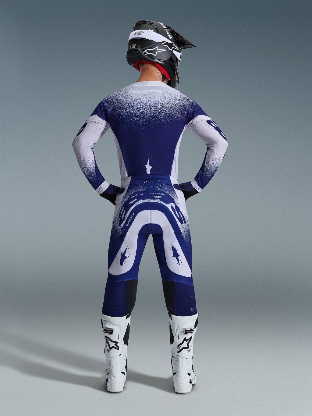 Alpinestars 2026 Supertech Scenz Pants, spodnie motocrossowe, jasnoszare i ciemnoniebieskie, widok z tyłu motocyklisty w pasującym zestawie odzieży z nakrapianą grafiką z efektem gradientu, białe buty i czarny kask, profesjonalny krój sportowy do wyścigów terenowych