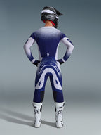 Alpinestars 2026 Supertech Scenz Pants, spodnie motocrossowe, jasnoszare i ciemnoniebieskie, widok z tyłu motocyklisty w pasującym zestawie odzieży z nakrapianą grafiką z efektem gradientu, białe buty i czarny kask, profesjonalny krój sportowy do wyścigów terenowych
