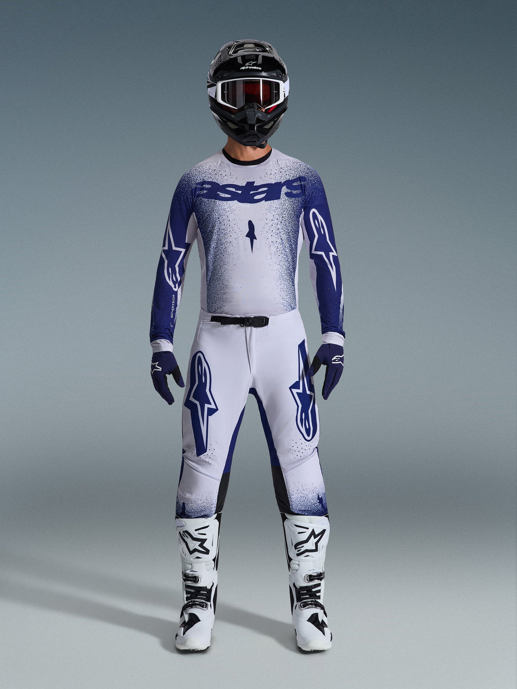 Motocyklista motocrossowy ubrany w zestaw odzieży Alpinestars 2026 Supertech Scenz, jasnoszary i ciemnoniebieski, zawierający koszulkę i spodnie z dopasowanymi niebieskimi rękawicami, biało-czarne buty oraz czarny kask z białymi goglami, skoordynowany strój do profesjonalnych wyścigów terenowych