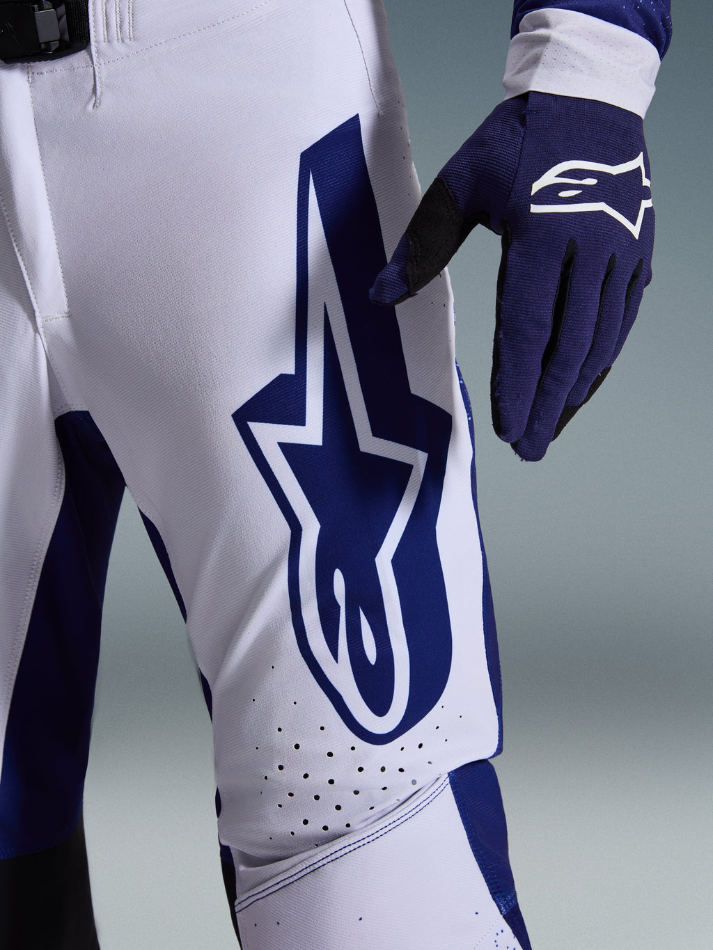 Spodnie Alpinestars 2026 Supertech Scenz, spodnie motocrossowe, jasnoszare i ciemnoniebieskie, zbliżenie na udo ukazujące duże ciemnoniebieskie logo Alpinestars, perforowane strefy wentylacyjne i elastyczny materiał rip-stop, noszone z pasującą ciemnoniebieską rękawicą motocrossową