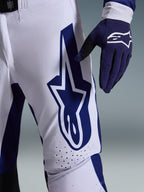 Spodnie Alpinestars 2026 Supertech Scenz, spodnie motocrossowe, jasnoszare i ciemnoniebieskie, zbliżenie na udo ukazujące duże ciemnoniebieskie logo Alpinestars, perforowane strefy wentylacyjne i elastyczny materiał rip-stop, noszone z pasującą ciemnoniebieską rękawicą motocrossową