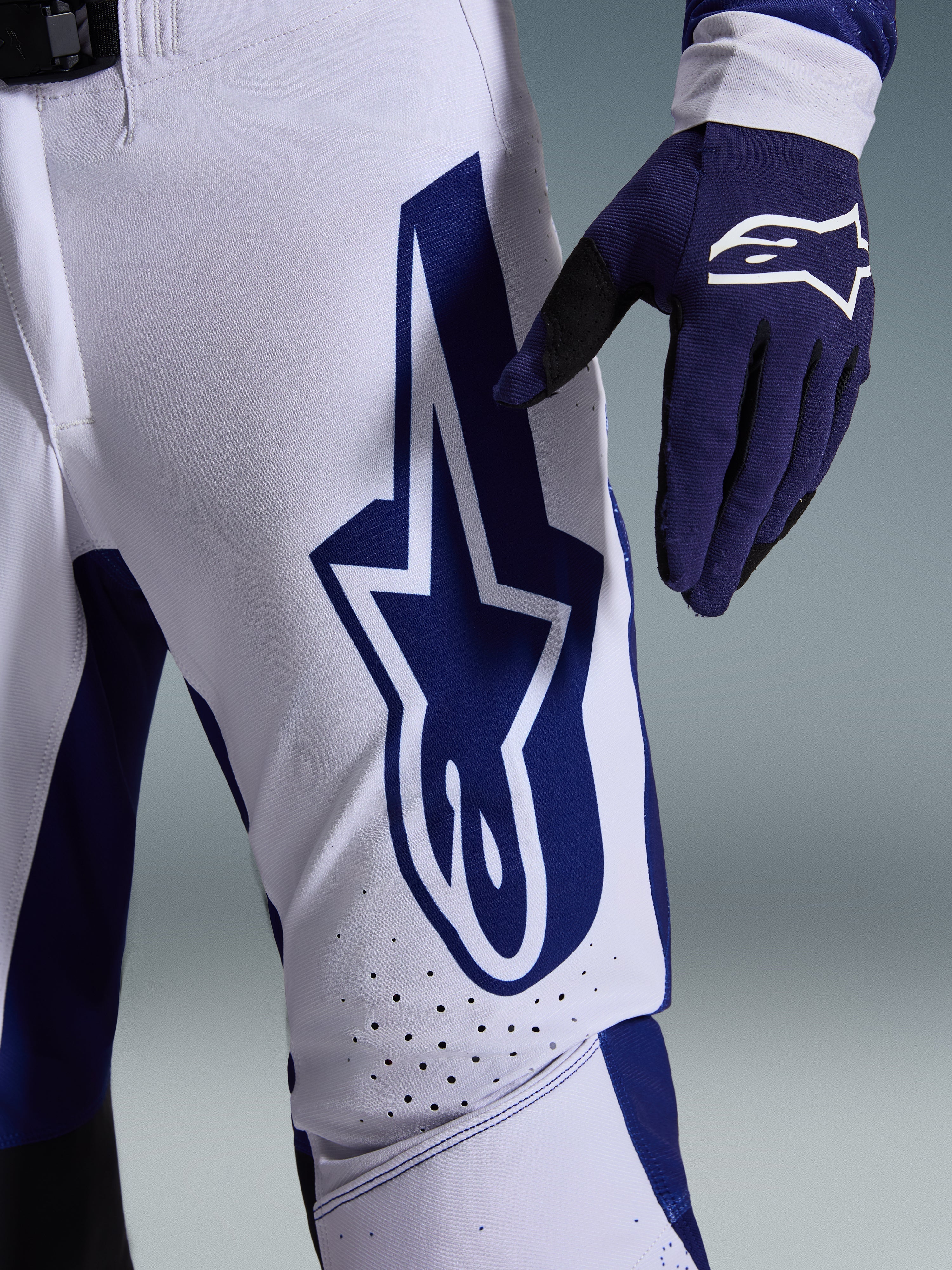 Spodnie Alpinestars 2026 Supertech Scenz, spodnie motocrossowe, jasnoszare i ciemnoniebieskie, zbliżenie na udo ukazujące duże ciemnoniebieskie logo Alpinestars, perforowane strefy wentylacyjne i elastyczny materiał rip-stop, noszone z pasującą ciemnoniebieską rękawicą motocrossową