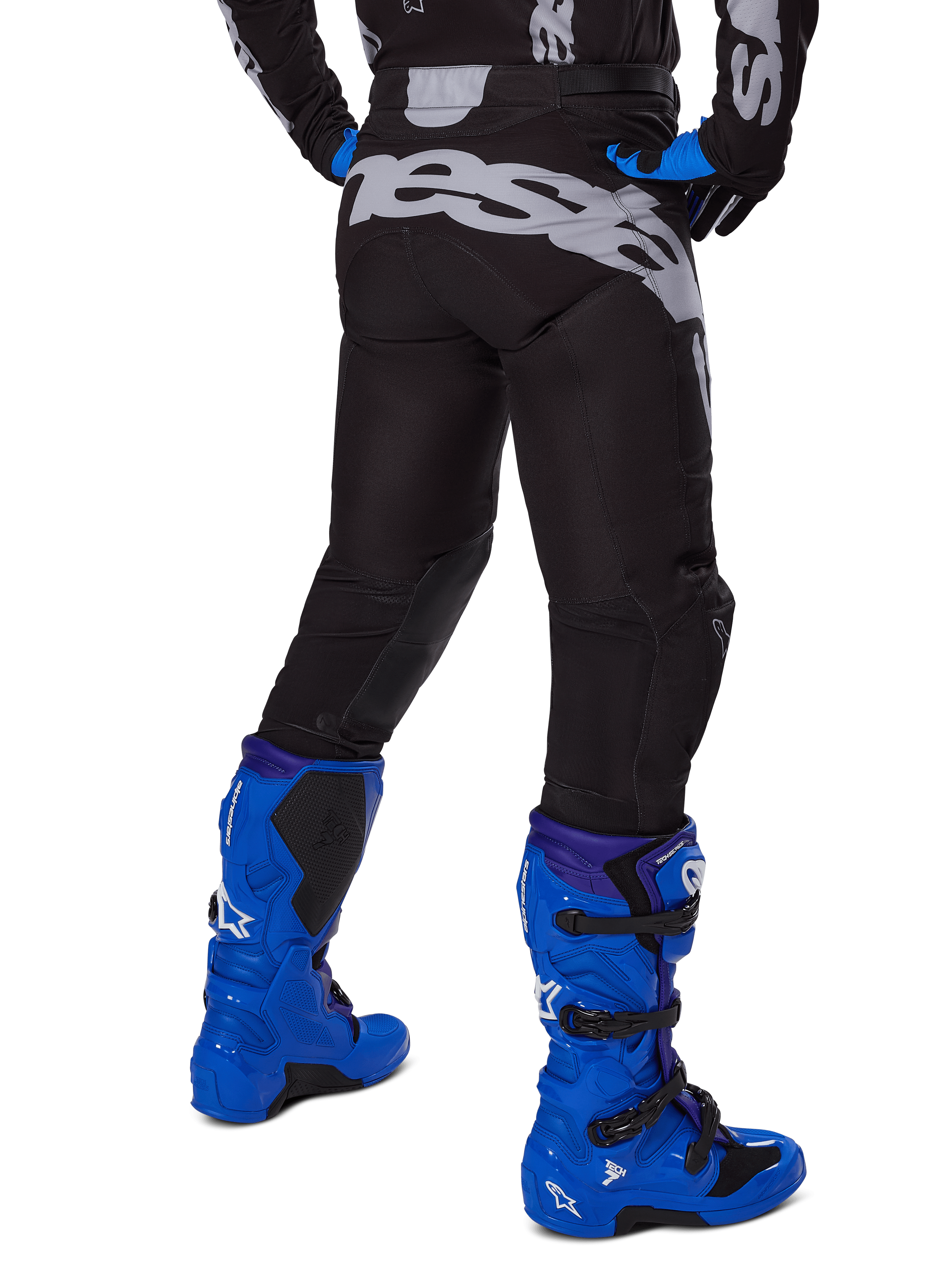 Alpinestars 2025 Racer Graphite Pants, spodnie motocrossowe, czarno-szare, widok z tyłu z dużym szarym brandingiem na siedzeniu, zestawione z jaskrawoniebieskimi butami motocrossowymi i czarno-niebieską koszulką, wytrzymała odzież off-road